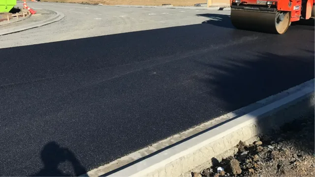 Roller compacting asphalt beside a concrete kerb and prepared pavement base.
