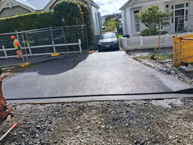 Freshly placed concrete driveway between residential houses, with temporary fencing and prepared aggregate in the foreground.
